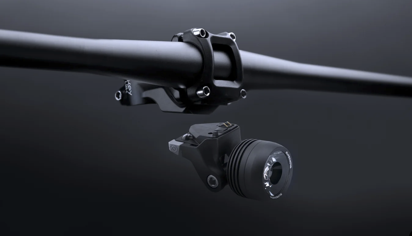 Supernova Mini 2 Fahrradscheinwerfer mit MonkeyLink Aufnahme in schwarz, schwebend unter einem LSupernova Mini 2 Fahrradscheinwerfer mit MonkeyLink Aufnahme in schwarz, schwebend unter einem Lenker, alles in grau schwarz gehalten