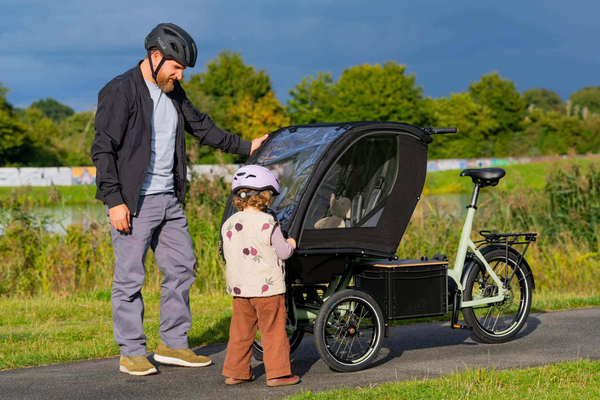 Ein Erwachsener steht auf einem Weg neben einem Chike e‑Kids Lastenrad mit Kinderkabine, in der ein kleines Kind steht. Beide tragen Fahrradhelme, im Hintergrund Wiese und Bäume.